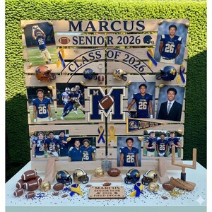 May include: A wooden display board with photos, footballs, and miniature helmets. The board features the text "MARCUS SENIOR 2026" and "CLASS OF 2026". Decorative elements include blue and gold ribbons and a miniature goalpost.