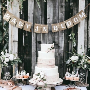 Op de afbeelding: Een witte vierlagen bruidstaart met een rustieke houten achtergrond en een banner van jute met de tekst "Baby in Bloom".