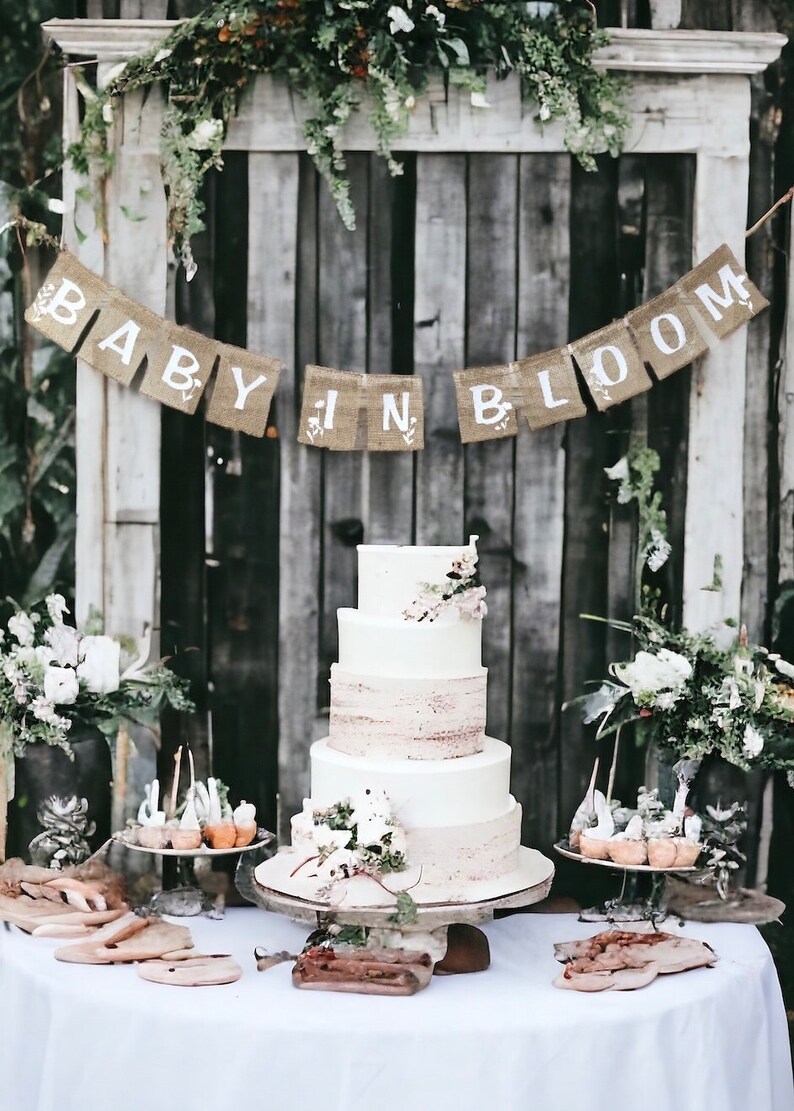 Op de afbeelding: Een witte vierlagen bruidstaart met een rustieke houten achtergrond en een banner van jute met de tekst "Baby in Bloom".