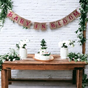Op de afbeelding: Een rustieke houten tafel met een witte bakstenen muur als achtergrond. Een banner van jute met de woorden "Baby in Bloom" hangt boven de tafel. Twee witte vazen met witte bloemen en een drielaagse taart versierd met groen staan op de tafel.