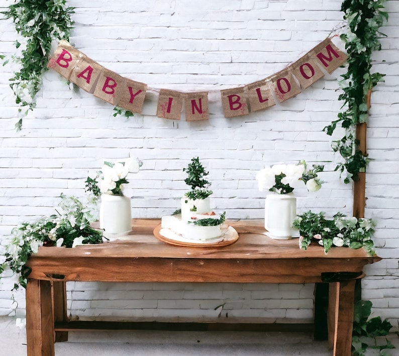 Op de afbeelding: Een rustieke houten tafel met een witte bakstenen muur als achtergrond. Een banner van jute met de woorden "Baby in Bloom" hangt boven de tafel. Twee witte vazen met witte bloemen en een drielaagse taart versierd met groen staan op de tafel.
