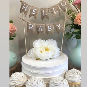 K&ouml;nnte beinhalten: Ein wei&szlig;er Kuchen mit einem Burlap-Banner, auf dem "Hello Baby" steht. Der Kuchen ist mit einer wei&szlig;en Blume verziert und von vier Cupcakes mit wei&szlig;em Frosting umgeben.