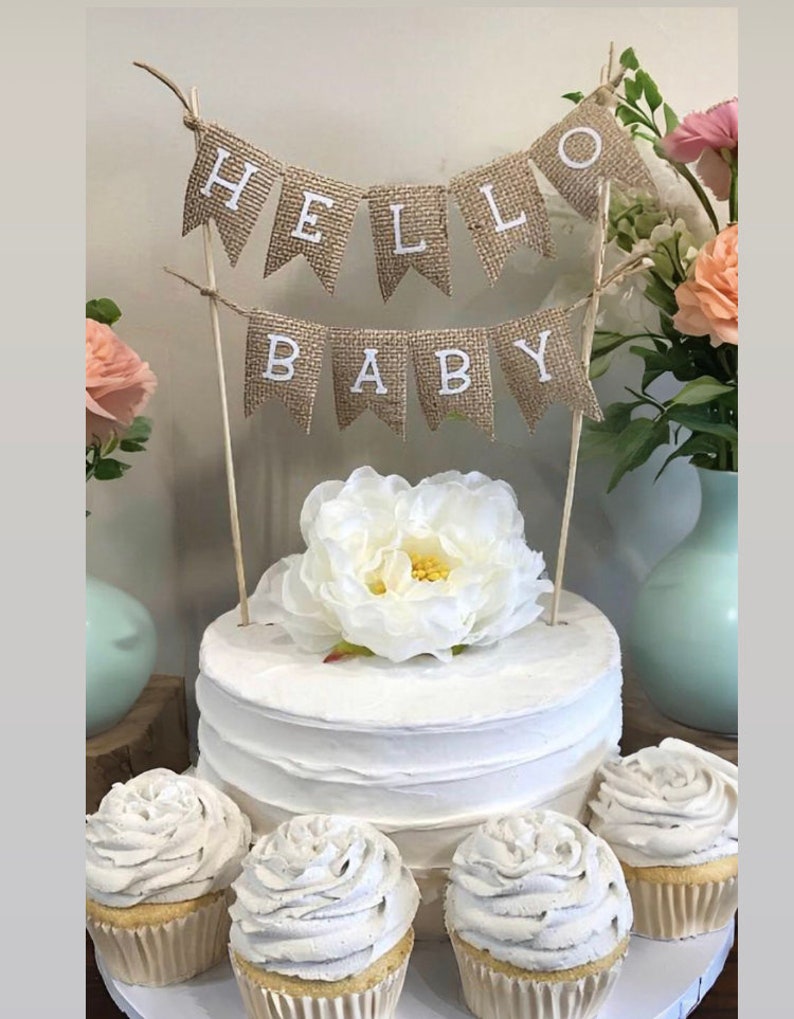 K&ouml;nnte beinhalten: Ein wei&szlig;er Kuchen mit einem Burlap-Banner, auf dem "Hello Baby" steht. Der Kuchen ist mit einer wei&szlig;en Blume verziert und von vier Cupcakes mit wei&szlig;em Frosting umgeben.