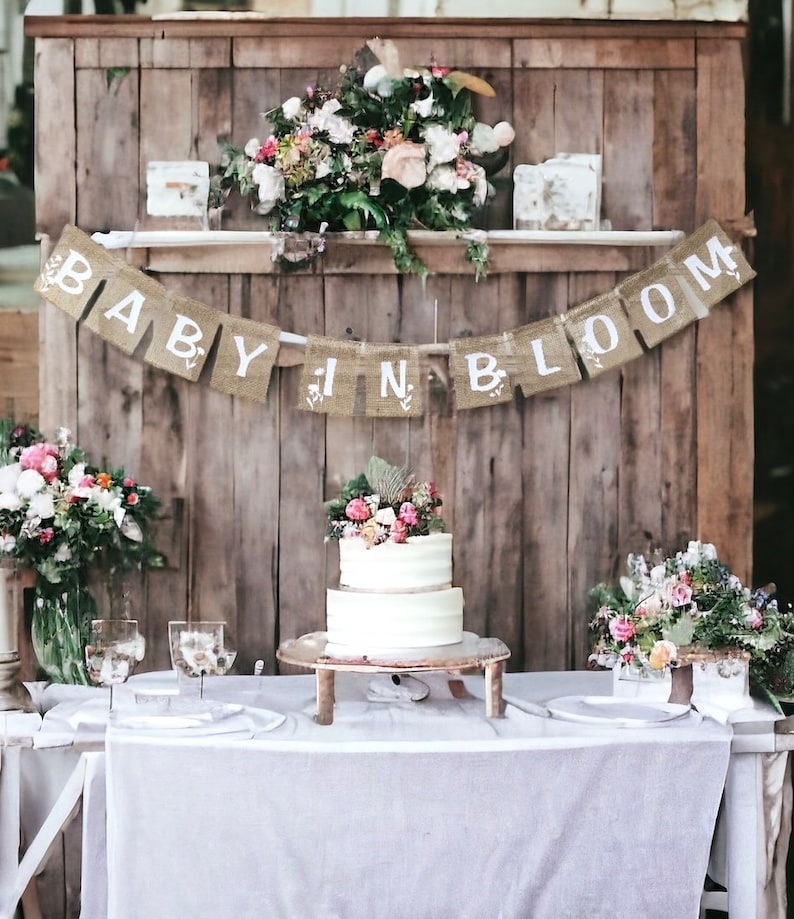 Op de afbeelding: Een rustieke houten achtergrond met een banner van jute die "Baby in Bloom" zegt. Een witte tweelaagse taart is versierd met verse bloemen en staat op een houten taartstandaard. De tafel is gedekt met witte tafelkleden en twee heldere glazen kelken.