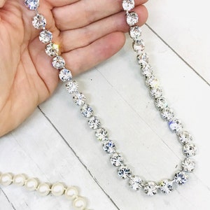 May include: A silver chain necklace with clear crystal stones. The necklace is on a white wooden surface.