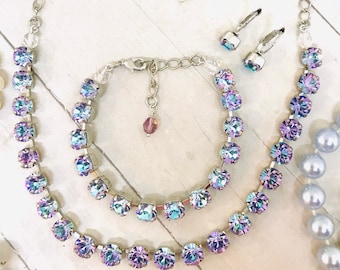 May include: A silver necklace, bracelet, and earrings set with iridescent purple and blue crystals. The necklace has two rows of crystals and a silver chain. The bracelet has one row of crystals and a silver clasp. The earrings are silver with a single crystal each.