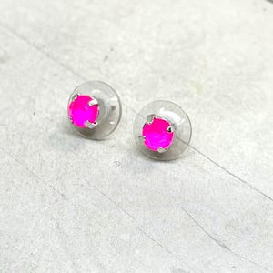 May include: A pair of stud earrings with bright pink, round gemstones set in silver-colored metal. The earrings are on clear, circular backings, and are displayed on a light gray surface.