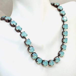 Peut inclure: Un collier délicat orné de pierres précieuses carrées bleu clair serties dans une monture en métal foncé. Le collier est présenté sur une surface blanche, mettant en valeur la couleur subtile des pierres et le travail du métal.