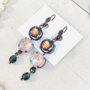 May include: Dangle earrings featuring copper-toned leverback hooks, each holding a circular cabochon with a sun graphic, a faceted iridescent crystal, and a small dark green bead at the bottom. The metal components have a dark copper finish.
