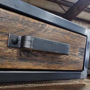 May include: Close-up of a drawer with a rustic wooden front and a black metal handle. The drawer is part of a larger piece of furniture with a black metal frame.