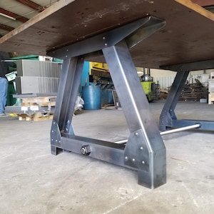 May include: A sturdy, industrial-style table with a large, rectangular wooden top and a metal base. The base is made of thick, black metal and has a triangular shape. The table is in a workshop setting.