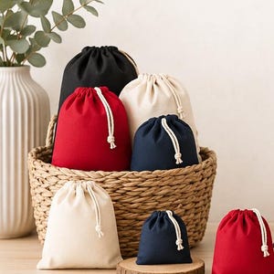 May include: A selection of drawstring bags in colours such as red, black, navy, and cream, are arranged in a woven basket. The bags have white drawstrings and are displayed on a wooden surface.