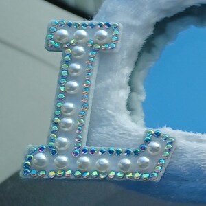 Op de afbeelding: Een witte letter L met een rand van parels en regenboogkleurige strass.