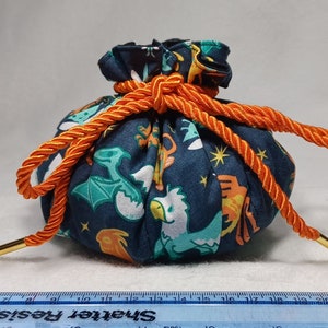 May include: A blue fabric drawstring bag with a colorful print of mythical creatures. The bag is tied with a bright orange rope and has a gold metal drawstring.