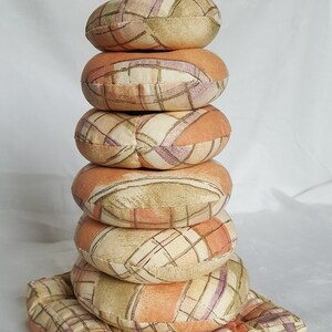 May include: A stack of six round, patterned cushions in shades of peach, beige, and lavender. The cushions are arranged in a tower, with a square base. The fabric has a grid-like design.