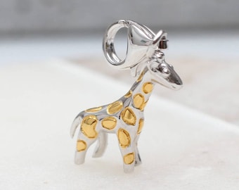 May include: A silver-toned giraffe charm featuring a polished finish with gold-toned spots across its body. The charm includes a circular bail at the top for attachment, showcasing a detailed animal figure with a smooth, reflective surface.