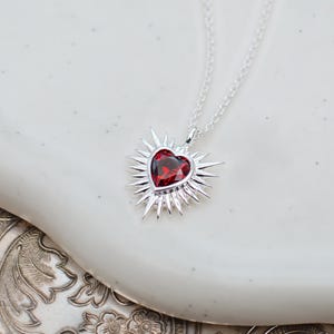 May include: A silver necklace featuring a heart-shaped pendant with a faceted red gemstone at the centre. The heart is surrounded by a sunburst design with radiating silver spikes. The pendant hangs from a delicate silver chain link necklace.