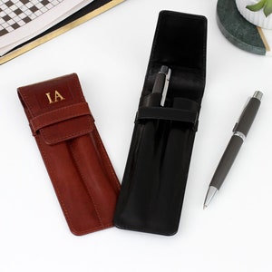 May include: Two leather pen cases, one in brown and one in black, with a pen in the black case and another beside it. The brown case features the gold initials "IA". The cases are rectangular with a flap closure.