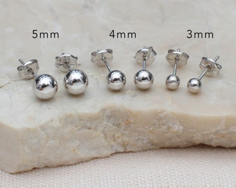 May include: Three pairs of silver-toned ball stud earrings with butterfly backs, arranged by size. The pairs are labeled 5mm, 4mm, and 3mm, showing a progression from the largest to the smallest spheres. Each earring features a polished, reflective surface.