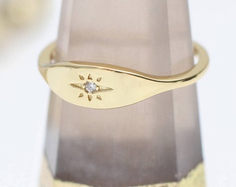 May include: A gold ring featuring a smooth, polished band with an oval-shaped face. The center of the face is engraved with a starburst pattern containing a single, small, clear, round-cut gemstone set in the middle of the design.