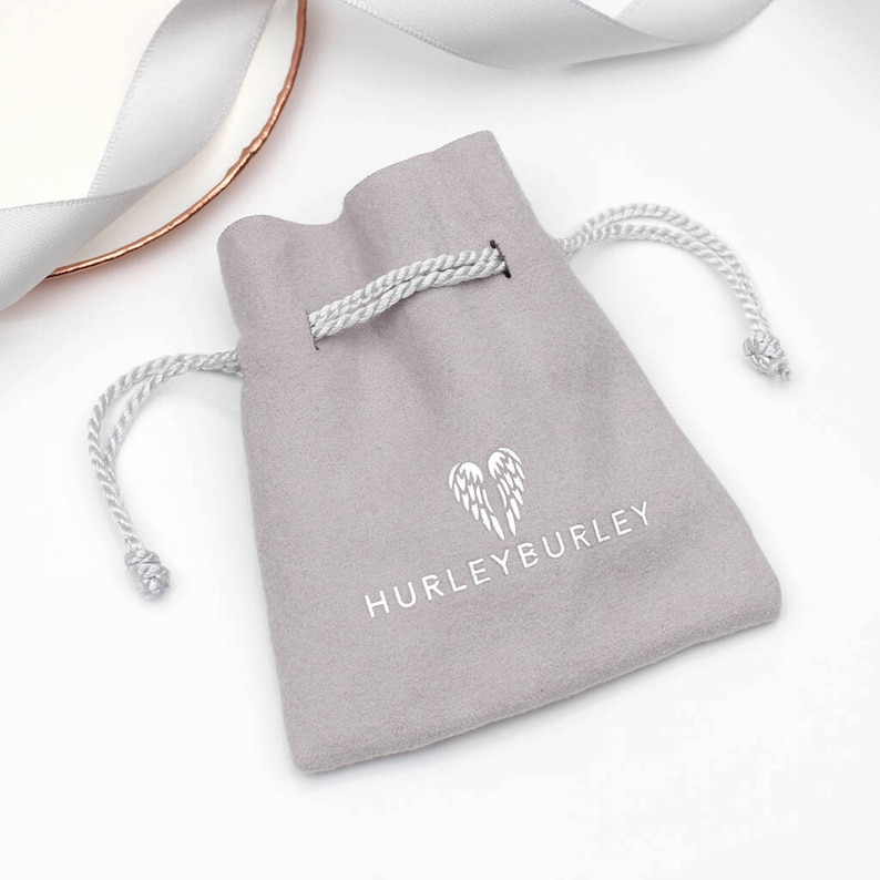 May include: A grey velvet drawstring pouch with the Hurleyburley logo printed in white. The logo features a pair of angel wings inside a heart.