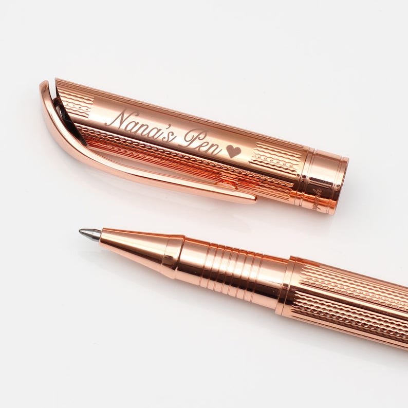 May include: Rose gold pen with the inscription "Nana's Pen" and a heart symbol. The pen's cap is open, revealing the pen's tip. The pen has a textured design on the barrel and cap.