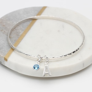 May include: A silver bangle bracelet with a hammered finish and two charms. One charm is a small blue gemstone and the other is a silver letter A.