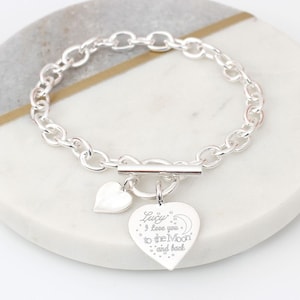 May include: Silver chain bracelet with a heart charm. The charm is engraved with the text "Lucy I love you to the Moon and back".