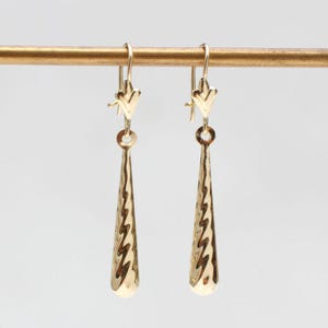 May include: A pair of gold drop earrings featuring a long, slender, teardrop-shaped pendant with a textured, wavy surface pattern. The earrings are suspended from simple gold hooks, showcasing a polished metallic finish that reflects light.
