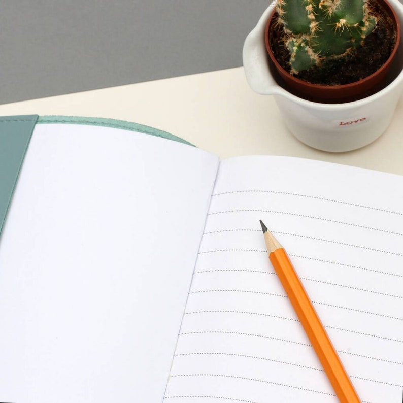 Puede incluir: Un cuaderno abierto con l&iacute;neas y una cubierta de color verde azulado con un l&aacute;piz amarillo en la p&aacute;gina derecha. El cuaderno est&aacute; sobre una superficie beige junto a una maceta de cer&aacute;mica blanca con una planta de cactus y la palabra "Love" escrita en el lateral.