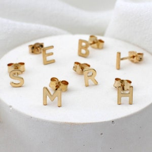 May include: Gold-coloured initial stud earrings. The earrings are in the shape of the letters S, E, B, R, M, H, and I. The earrings are displayed on a white surface.