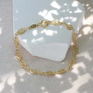 May include: A gold chain bracelet featuring a series of repeating oval-shaped links with intricate filigree scrollwork patterns. The bracelet is secured with a round spring ring clasp and includes a small rectangular metal tag near the closure.