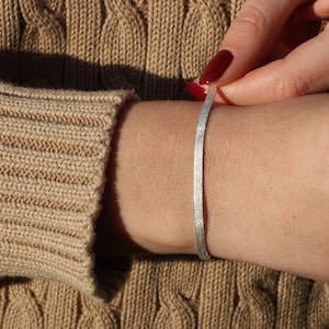 May include: A silver bracelet with a textured finish. The bracelet is thin and delicate, and it is shown on a person's wrist.