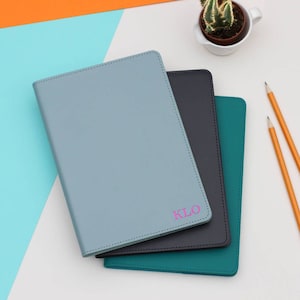 Puede incluir: Tres cuadernos encuadernados en cuero de color azul, verde azulado y negro. El cuaderno superior tiene el nombre "KLO" impreso en rosa.