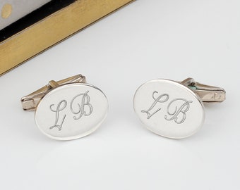 May include: A pair of silver oval cufflinks. Each cufflink features the engraved initials "LB" in an elegant script. The cufflinks have a polished finish and a classic design, suitable for formal wear.