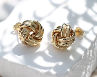 May include: A pair of gold stud earrings featuring a polished, knotted design. Each earring consists of intertwined metallic loops forming a spherical shape, complete with standard post backings for secure attachment to the earlobe.