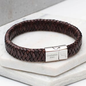 Op de afbeelding: Een donkerbruine gevlochten leren armband met een gepolijste zilverkleurige metalen sluiting. De sluiting is gegraveerd met de tekst Graduation 2021 en een klein icoontje van een afstudeerhoed. De armband heeft een ronde vorm en textuur.