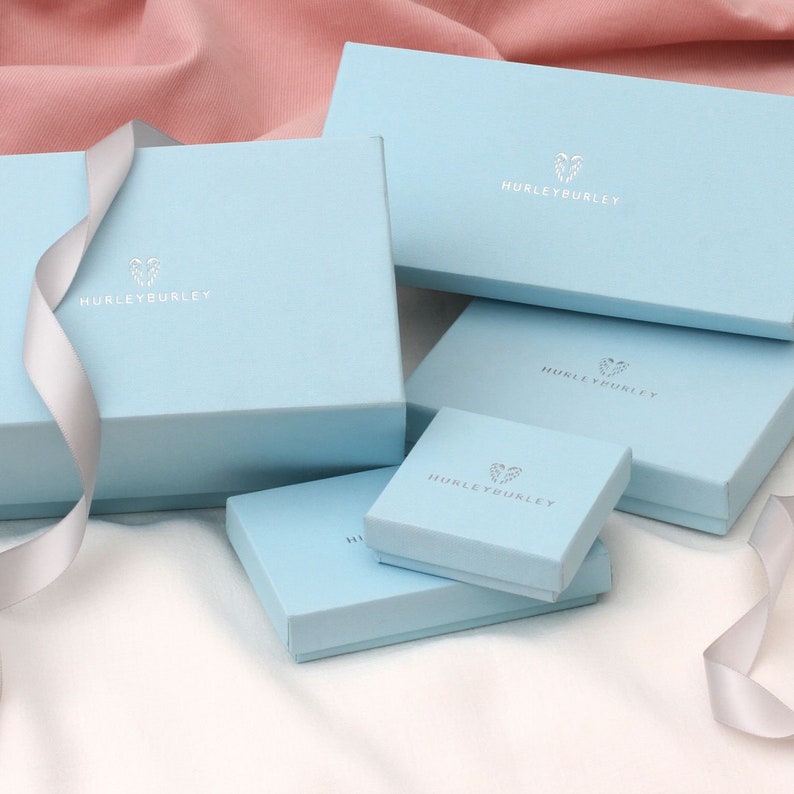 May include: A collection of light blue gift boxes in various sizes, some stacked, with the brand name "HURLEYBURLEY" printed on them. A silver ribbon is draped across the boxes. The boxes are on a white surface with a pink background.
