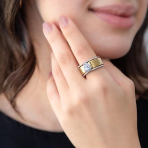Może przedstawiać: Szeroki pierścionek z polerowanym złotym środkiem otoczonym dwoma srebrnymi, teksturowanymi krawędziami. Pojedynczy, przezroczysty kamień w kształcie gruszki jest osadzony na obrączce, otoczony aureolą małych, błyszczących białych kamieni.