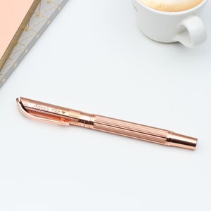 May include: Rose gold pen with the inscription "Nana's Pen" and a heart symbol. The pen has a textured, patterned design on the barrel and a smooth, curved clip. A cup of coffee and a notebook are in the background.