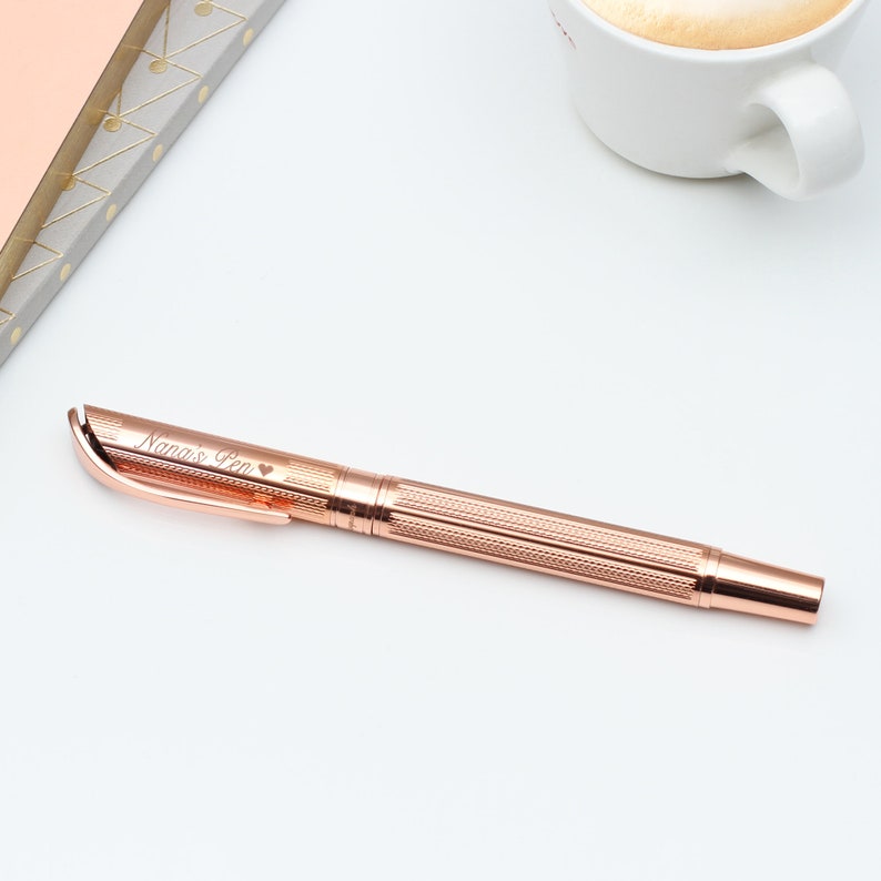 May include: Rose gold pen with the inscription "Nana's Pen" and a heart symbol. The pen has a textured, patterned design on the barrel and a smooth, curved clip. A cup of coffee and a notebook are in the background.