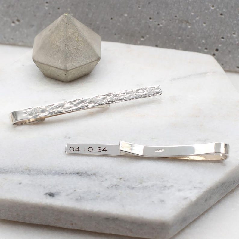 May include: Silver tie bar with a textured surface and engraved with the date "04.10.24".