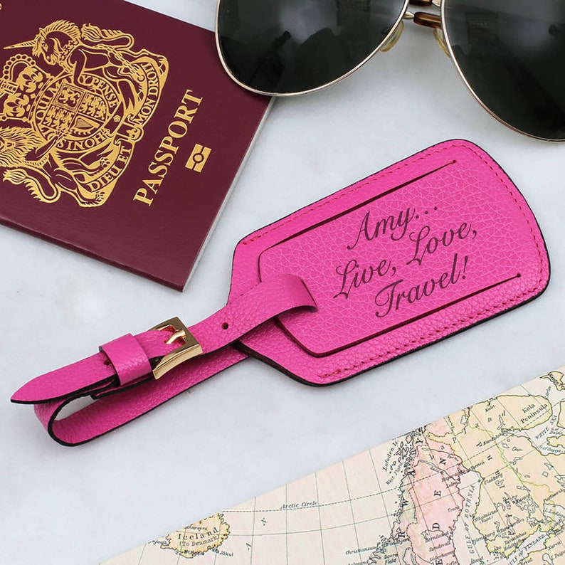 May include: A bright pink luggage tag with the text "Amy... Live, Love, Travel!" is displayed next to a passport and sunglasses. The tag has a gold buckle and strap. A map and sunglasses are also in the image.