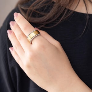 May include: A wide band ring featuring a polished gold-tone center section flanked by two silver-tone twisted rope-style edges. The ring has a substantial profile and a two-tone metallic finish, showcasing a smooth central band with textured borders.