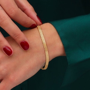 Könnte beinhalten: Ein goldfarbenes Armband mit gewebtem Design. Das Armband wird an einem Handgelenk mit rotem Nagellack getragen. Der Hintergrund ist ein dunkelgrüner Stoff.