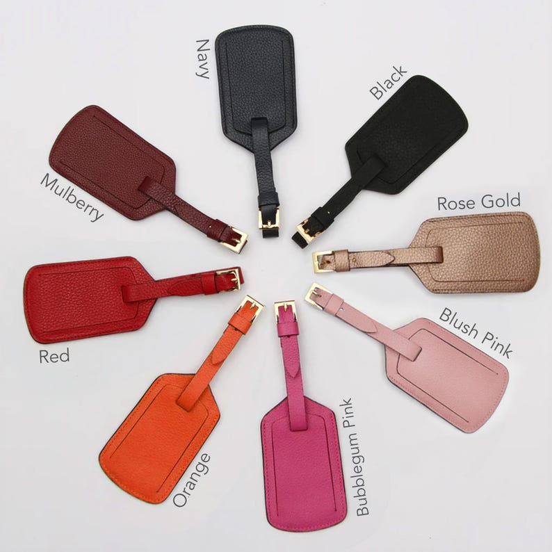 May include: A collection of leather luggage tags in various colors, including red, orange, pink, rose gold, black, and navy. Each tag has a rectangular shape with a strap and a gold-colored buckle. The tags are arranged in a circular pattern.