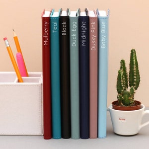 Puede incluir: Siete cuadernos de diferentes colores con los siguientes colores: mora, verde azulado, negro, huevo de pato, medianoche, rosa polvoriento y azul beb&eacute;. Los cuadernos est&aacute;n alineados en una fila sobre una mesa.