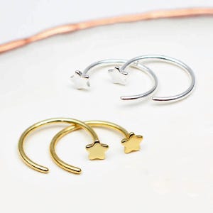 Op de afbeelding: Twee paar open oorringen, één paar in zilver en één paar in goud. Elke oorbel is voorzien van een kleine, vijfpuntige sterbedel bevestigd aan één uiteinde van de dunne, cirkelvormige draadring. De oorbellen zijn gerangschikt in twee sets.