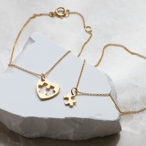 May include: Two gold necklaces with puzzle piece pendants. One necklace has a heart-shaped pendant with two puzzle pieces inside. The other necklace has a single puzzle piece pendant.