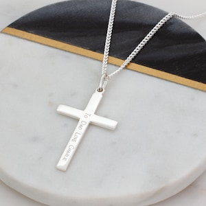 Puede incluir: Un colgante de cruz de plata en una cadena de plata a juego. La barra vertical de la cruz está grabada con el texto 'To Dad Love Charlie' en una fuente sans-serif clara. El colgante presenta un acabado metálico pulido y reflectante.